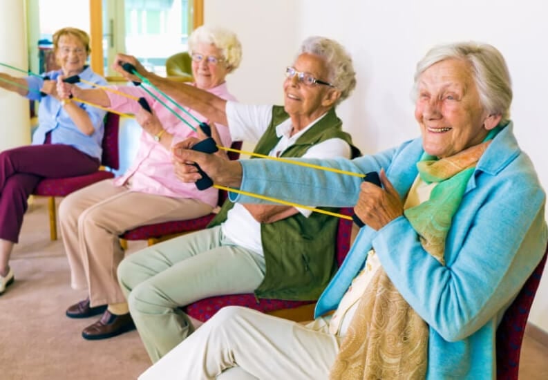 Elderly Couple Exercising On Medicine Balls Inside Memory Care Oklahoma City Elderly Residents Participating in Group Exercise Class With Resistance Bands at Memory Care